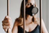 Молодая женщина в защитной респираторной маске и черном платье стоит за металлическим забором и смотрит в сторону, представляя концепцию коронавирусной профилактики и изоляции — стоковое фото