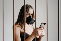 Молодая женщина в черной респираторной маске для предотвращения коронавируса стоит за решеткой и просматривает мобильный телефон — стоковое фото