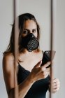 Mujer joven con máscara respiratoria negra para la prevención del coronavirus de pie detrás de las rejas y el teléfono móvil de navegación - foto de stock