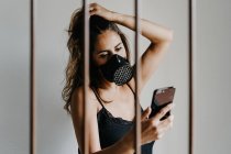 Молодая женщина в черной респираторной маске для предотвращения коронавируса стоит за решеткой и просматривает мобильный телефон — стоковое фото