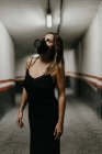 Веселая молодая женщина в элегантном черном платье и черной респираторной маске смотрит вверх, стоя в узком коридоре внутри здания — стоковое фото