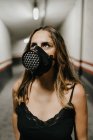 Jovem mulher alegre em vestido preto elegante e máscara respirador preto olhando para cima, enquanto está em pé no corredor estreito dentro do edifício — Fotografia de Stock