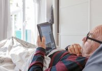 Vue latérale d'un homme âgé portant des lunettes couché au lit et lisant un magazine électronique sur tablette pendant qu'il passait la matinée à la maison — Photo de stock