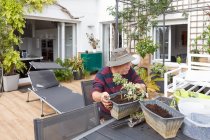 Seitenansicht eines Rentners in Freizeitkleidung und Hut, der Setzlinge im Topf pflanzt, während er am Tisch im Garten neben dem Haus sitzt — Stockfoto