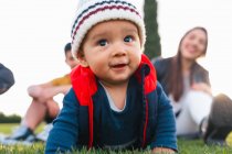 Nettes ethnisches Kleinkind in warmer Kleidung lächelt beim Krabbeln auf der grünen Wiese während des Familienwochenendes in der Natur — Stockfoto