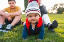 Lindo niño étnico en ropa de abrigo sonriendo y mirando a la cámara mientras se arrastra en el prado verde durante el fin de semana familiar en el campo - foto de stock
