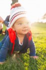 Cute ethnic toddler in warm clothes smiling while crawling on green meadow during family weekend in countryside — Stock Photo