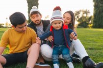 Cheerful young man and woman with teen boy and toddler kid smiling and looking at camera while sitting on plaid on green grass and enjoying time together in sunny autumn day in park — Stock Photo