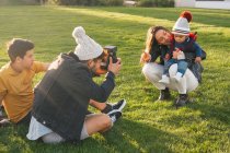 Jovem com câmera instantânea sentada na grama perto do filho adolescente e tirando foto de esposa alegre com criança pequena enquanto passam o tempo juntos no parque de outono — Fotografia de Stock
