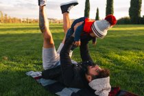 Vista laterale del giovane allegro sdraiato su una coperta sull'erba verde e con in braccio un bambino carino in caldo mentre riposa durante il fine settimana nel parco — Foto stock