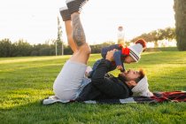 Side view of cheerful young man lying on blanket on green grass and holding cute toddler kid in warm wear while resting during weekend in park — Stock Photo