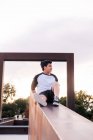 Basso angolo di positivo giovane uomo in abbigliamento casual seduto sulla recinzione di pietra e distogliendo lo sguardo mentre riposa nel parco cittadino in serata di sole — Foto stock