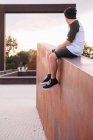 Low angle of positive young man in casual wear sitting on stone fence and looking away while resting in city park in sunny evening — Stock Photo