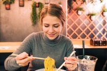 Молода самка, сидячи за столом у японському ресторані, паличками й ложкою, їсть смачні рамена. — стокове фото