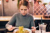 Jeune femme utilisant des baguettes et une cuillère pour manger de savoureux ramen assis à table dans un restaurant japonais — Photo de stock
