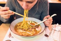 Молодая женщина, сидя за столом в японском ресторане, ест вкусный рамен палочками для еды и ложкой — стоковое фото