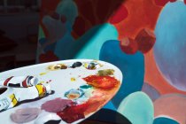 Paleta bagunçada com manchas de pigmento e tubos colocados perto do cavalete com pintura abstrata — Fotografia de Stock
