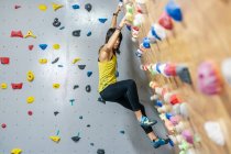 Side view from below of focused woman in sportswear hanging on steep wall above mats in modern climbing center — Stock Photo