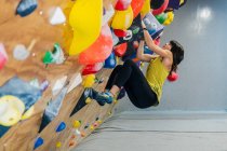Side view from below of focused woman in sportswear hanging on steep wall above mats in modern climbing center — Stock Photo