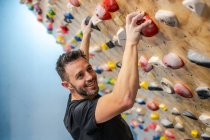Vista laterale di forte atleta maschio felice guardando la fotocamera in abbigliamento sportivo arrampicata su parete colorata durante l'allenamento in palestra moderna — Foto stock