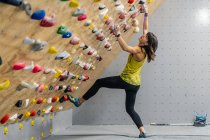 Vista lateral de baixo de mulher focada em sportswear pendurado na parede íngreme acima tapetes no moderno centro de escalada — Fotografia de Stock