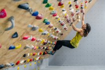Вид снизу сфокусированной женщины в спортивной одежде, висящей на крутой стене над ковриками в современном альпинистском центре — стоковое фото