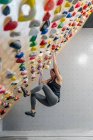 Vue latérale d'en bas de la femme concentrée en vêtements de sport suspendus sur un mur escarpé au-dessus des tapis dans le centre d'escalade moderne — Photo de stock