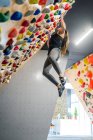 Вид збоку від зосередженої жінки в спортивному одязі, що звисає на крутій стіні над килимками в сучасному центрі скелелазіння — стокове фото