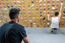 Vue arrière de l'athlète masculin anonyme en vêtements de sport regardant pour grimpeur pendant l'entraînement sur le mur dans la salle de gym — Photo de stock