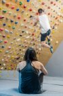 Back view of unrecognizable focused female in sportswear sitting on floor and watching male blurred anonymous climber on wall during workout in gym — Stock Photo