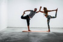 Seitenansicht eines ruhigen Paares in Sportbekleidung, das in Natarajasana auf einer Yogamatte steht, sich Händchen haltend und einander anschauend — Stockfoto