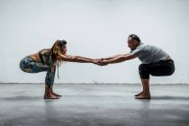 Vista lateral do casal foco em sportswear praticando ioga em Utkatasana enquanto estão descalços no chão e olhando um para o outro — Fotografia de Stock