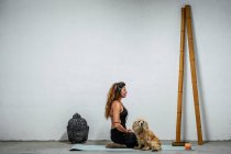 Vista lateral de contenido femenino sentado en esterilla de yoga con perro Cocker Spaniel inglés y meditando en Padmasana en habitación con cabeza de buda y palos de bambú - foto de stock
