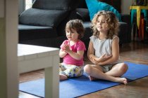 Positive kleine Schwestern, die mit ausgestreckten Armen in Lotus-Pose sitzen, während sie Video-Tutorial ansehen und zu Hause im Wohnzimmer gemeinsam Yoga praktizieren — Stockfoto