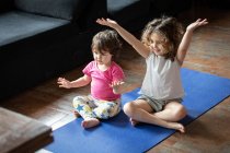 High angle of positive little sisters sitting in lotus pose with outstretched arms while watching video tutorial and practicing yoga together in living room at home — Stock Photo