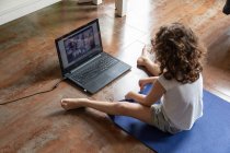 Назад вид неузнаваемой маленькой девочки, смотрящей онлайн видео учебник на ноутбуке, сидя на коврике и изучая йогу позировать дома — стоковое фото