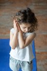 Ângulo alto de calma pequena menina de cabelos encaracolados em uso casual mantendo as mãos em gesto namaste enquanto estava em pé no tapete de ioga durante a aula de ioga em casa — Fotografia de Stock