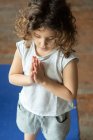 Angle élevé de calme petite fille aux cheveux bouclés en tenue décontractée gardant les mains en geste namaste tout en se tenant debout sur un tapis de yoga pendant un cours de yoga à la maison — Photo de stock