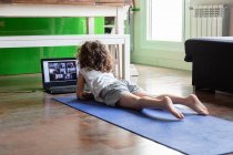 Back view of unrecognizable little girl watching online video tutorial on laptop while sitting on mat and learning yoga pose at home — Stock Photo