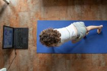 Rückansicht von unkenntlich kleinen Mädchen, die Online-Video-Tutorial auf Laptop anschauen, während sie auf Matte sitzen und zu Hause Yoga-Pose lernen — Stockfoto
