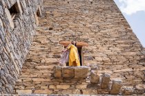 D'en bas de l'ancien bâtiment monumental avec une touriste féminine éloignée sortant de la porte en robe jaune tout en profitant d'une chaude journée ensoleillée dans le village de marbre à Al Bahah — Photo de stock