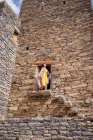 Desde abajo del monumental edificio antiguo con la remota turista femenina que sale de la puerta en vestido amarillo mientras disfruta de un día soleado caliente en Marble Village en Al Bahah - foto de stock