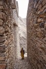 Віддалена жінка в барвистому одязі, що йде між обшарпаними стародавніми стінами історичних будинків села Марбл в Аль-Баха, розташованих на гірській місцевості в Саудівській Аравії. — стокове фото