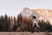 Visão lateral de corpo inteiro de jovem viajante despreocupado em roupas elegantes correndo e pulando no prado contra pinheiros e majestoso penhasco rochoso no dia ensolarado da primavera no Parque Nacional de Yosemite, nos EUA — Fotografia de Stock