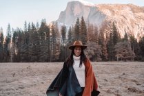 Happy relaxed young female traveler in stylish outfit sitting on stone border against picturesque mountain scenery with rocky cliffs and coniferous forest in Yosemite National Park in USA — Stock Photo