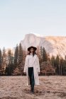 Heureuse jeune voyageuse détendue en tenue élégante assise sur la frontière de pierre contre des paysages de montagne pittoresques avec des falaises rocheuses et une forêt de conifères dans le parc national Yosemite aux États-Unis — Photo de stock