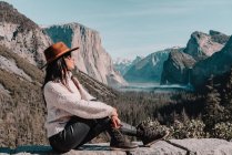 Full body side view of happy relaxed young female traveler in stylish outfit sitting on stone border against picturesque mountain scenery with rocky cliffs and coniferous forest in Yosemite National Park in USA — Stock Photo