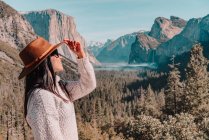 Full body side view of happy relaxed young female traveler in stylish outfit sitting on stone border against picturesque mountain scenery with rocky cliffs and coniferous forest in Yosemite National Park in USA — Stock Photo