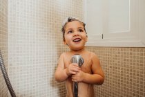 Усміхнена маленька дитина з піною на голові стоїть у ванній кімнаті з душем і співом, дивлячись вниз — стокове фото