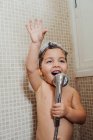 Улыбающийся маленький ребенок с пеной на голове стоит в ванной комнате с душем и поет, отводя взгляд — стоковое фото